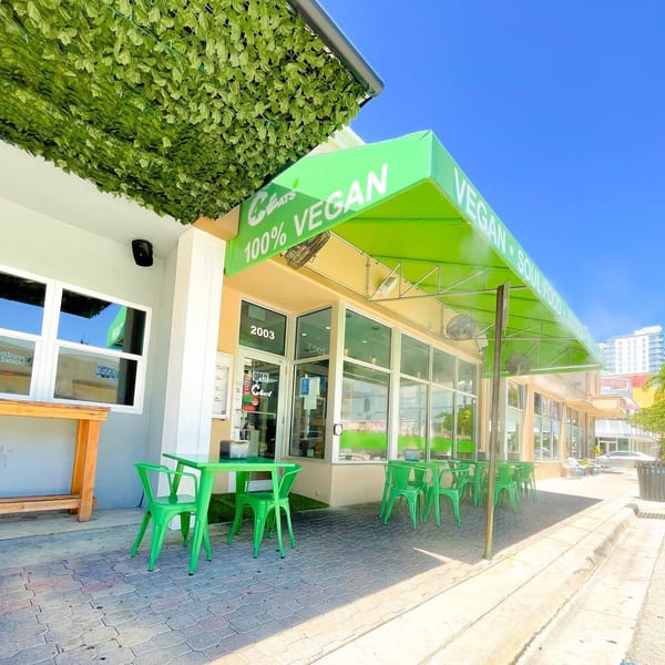 C4Eats vegan restaurant exterior with green awning, outdoor seating, and leafy decor under a blue sky.