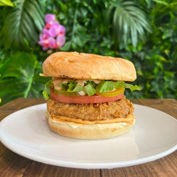 A vegan fried chicken sandwich with lettuce, tomato, pickles, and sauce on a white plate.