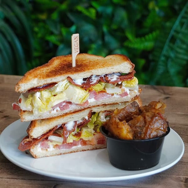 Vegan BLT sandwich with a "VEGAN" pick, served with a side of roasted potatoes.