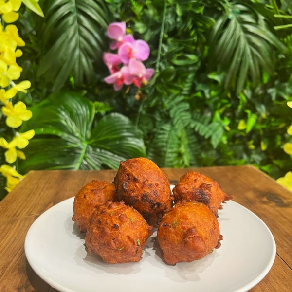 Five golden-brown vegan fritters on a white plate, with a lush green and floral background.